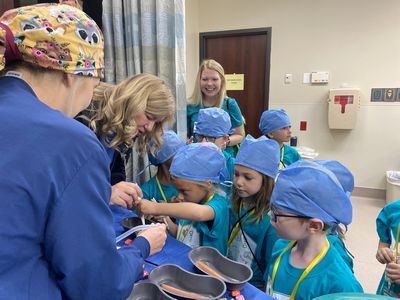 Children, wearing hospital scrubs, watch a medical professional demonstrate surgery on a model.