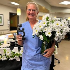 Daisy Award winner Cheryl Turner holds a vase of flowers and the award in front of a nurses station. 