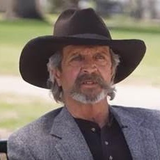 Emul King sits outside while wearing a black cowboy hat and a grey overcoat.