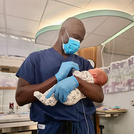 Dr. Patrick Christopher, wearing hospital scrubs and a blue face mask, holds Parker while feeding him a bottle.