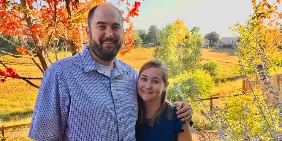 Katie and Jordan smile while standing together in front of a fall colors grassy field.