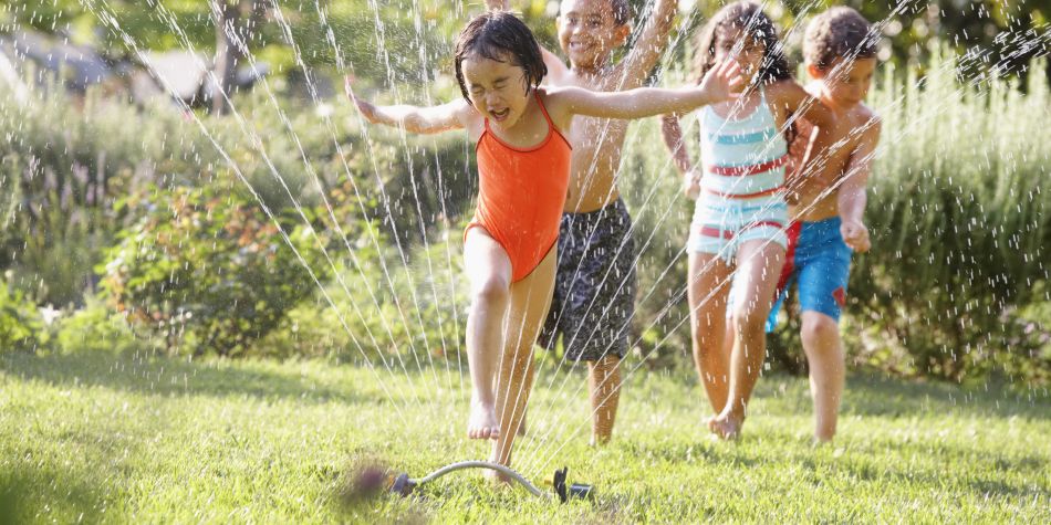 A group of children in a line are running on grass as they jump through a sprinkler.