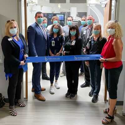 Hospital staff, wearing dress clothes and masks, stand together holding a large blue ribbon as they unveil the newly renovated Cardiac Catheterization Suite.